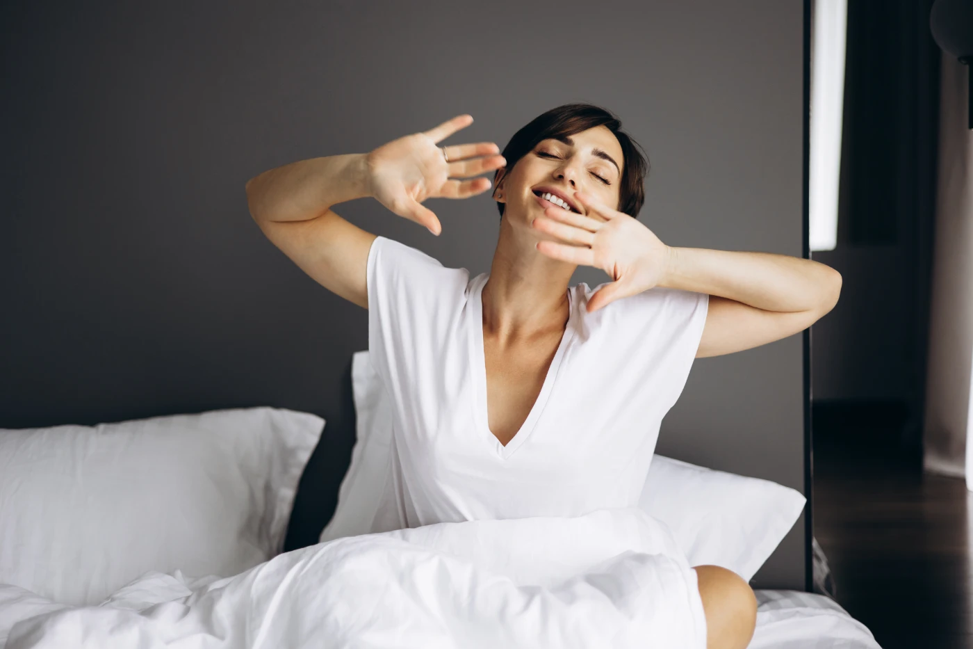 Woman stretches and smiles, sitting up in bed with white sheets and pillows.
