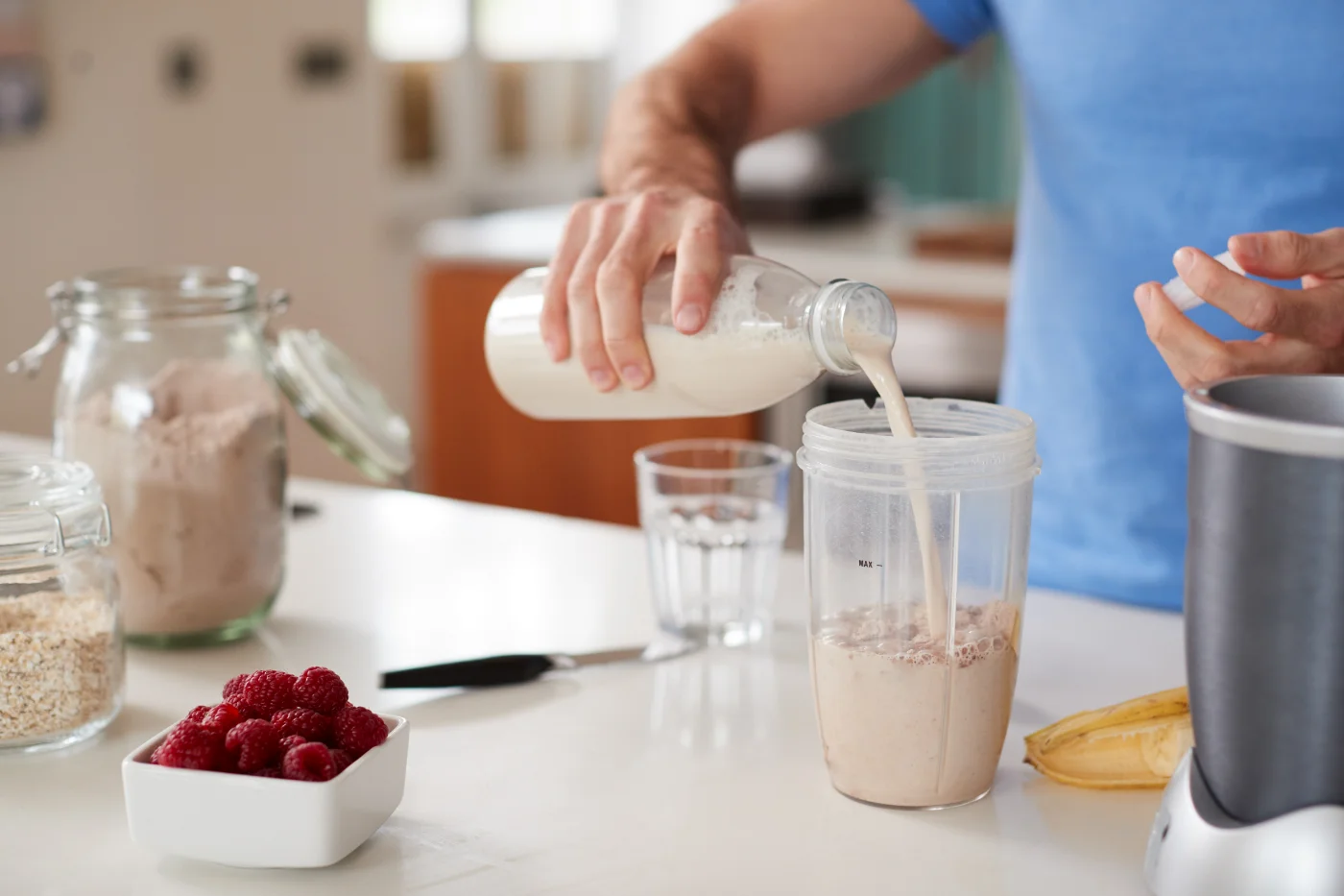 Person pouring milk into blender, preparing a smoothie with ingredients like raspberries, oats, and new zealand whey protein.