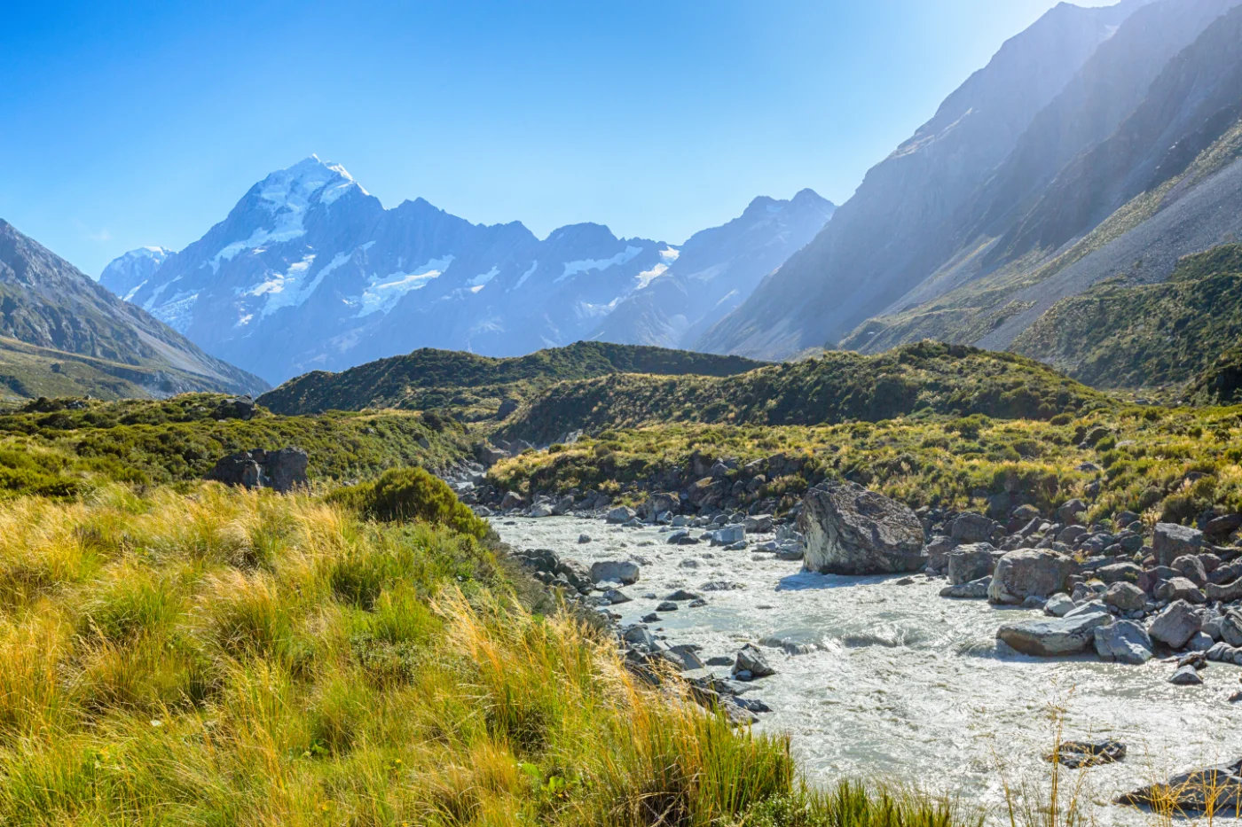 Mountain river flowing toward snow-capped peaks and green hills under a clear, blue sky.