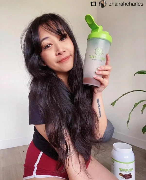 Woman holding a protein shaker; tub of protein powder visible nearby.