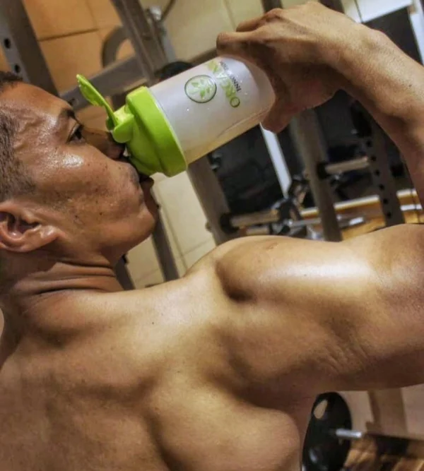 Muscular man drinking from a shaker bottle in a gym setting.