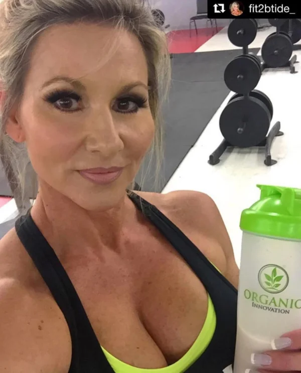 Woman in gym attire holding protein shake with weights in background.