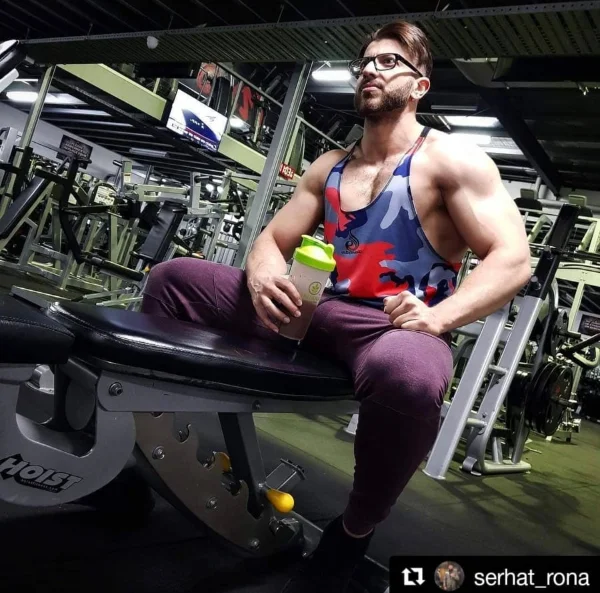 Muscular man with glasses and tank top sits on gym equipment, holding a protein shake.